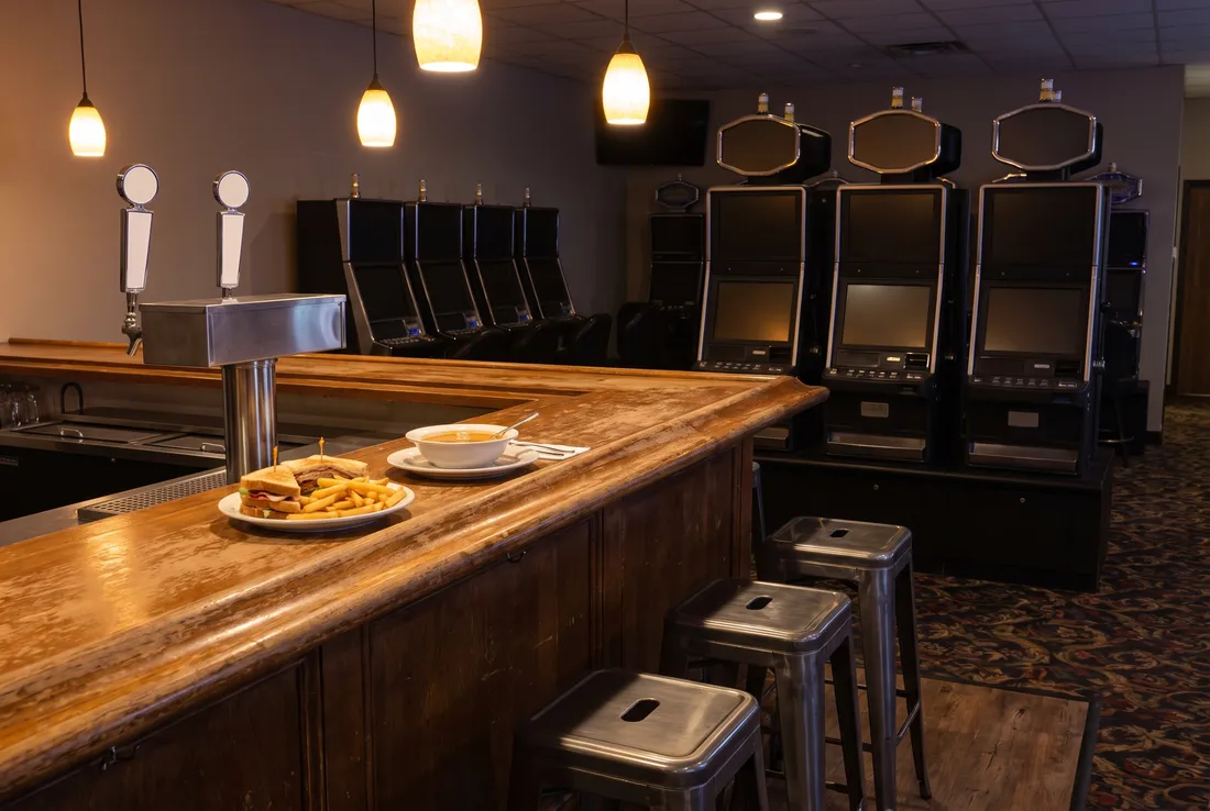 A short wooden bar counter with steel stools, a sandwich with fries and a bowl of soup, and slot machine cabinets with blank screens behind.
